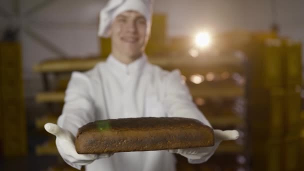 Feliz, jovem padeiro de uniforme apresenta pão de centeio para a câmera em uma padaria — Vídeo de Stock