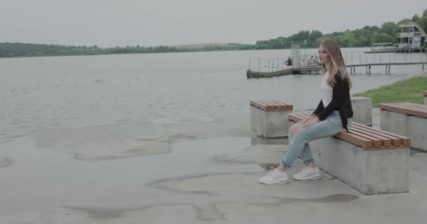 Κορίτσι που κάθεται στον πάγκο στην όχθη του ποταμού και θαυμάζοντας τα κύματα με τον άνεμο — Αρχείο Βίντεο