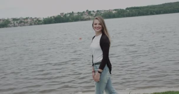 Fröhliches Mädchen in lässiger Kleidung, das bei Wind am Flussufer spaziert und posiert — Stockvideo