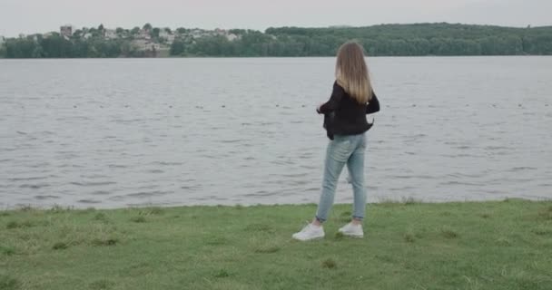 Rückansicht des Mädchens steht an einem windigen Tag am Fluss und korrigiert ihre Strickjacke — Stockvideo