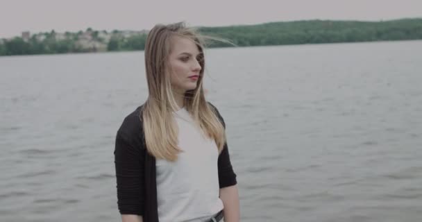 Jolie fille avec des cheveux soufflants regardant loin et tournant à la caméra à la rivière — Video