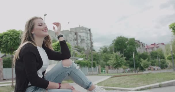 Girl with blowing hair and in casual clothes laughing and looks around on bench — 图库视频影像