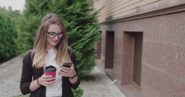 Biznes dziewczyna w okularach pije filiżankę kawy i za pomocą smartphoone na zewnątrz — Wideo stockowe