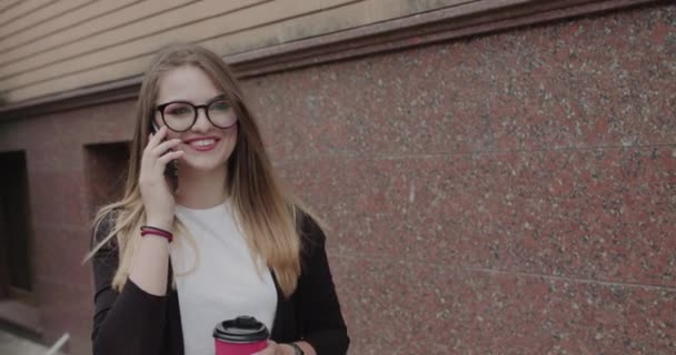 Chica alegre camina con taza de bebida en la mano y hablando por teléfono con sonrisa — Vídeo de stock
