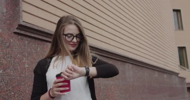 Uśmiechnięta dziewczyna w okularach trzyma filiżankę herbaty i patrzy na inteligentną bransoletkę na świeżym powietrzu — Wideo stockowe