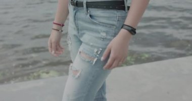 Pantolon giymiş bir kız nehir kenarında yürüyor ve elini cebine sokuyor.
