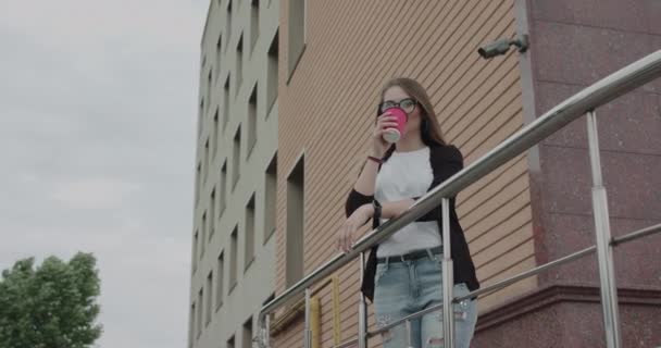 Счастливая девушка пьет бокал напитка и улыбается, опираясь на перила на улице. — стоковое видео
