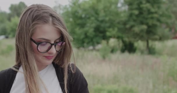 Дівчина з дме волоссям дивиться на камеру з посмішкою на обличчі і виправляє окуляри — стокове відео