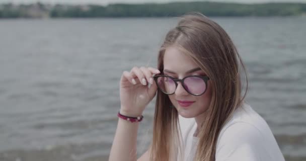 Chica bonita se relaja en la cubierta en el río en el día ventoso, sostiene las gafas y mira a un lado — Vídeo de stock