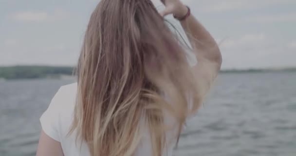 Mädchen korrigiert wehende Haare, wendet sich der Kamera zu und lächelt bei Wind am Fluss — Stockvideo