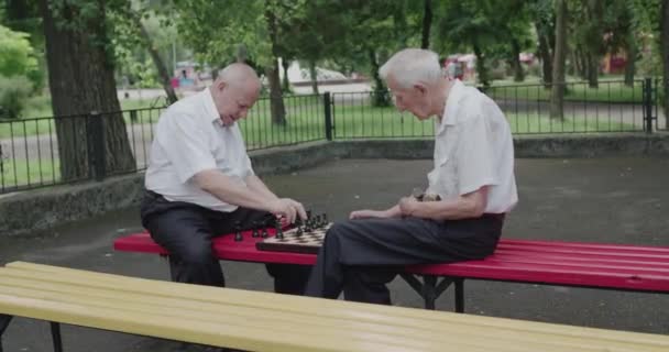 Двоє літніх друзів грають у шахи на вуличній лавці влітку — стокове відео