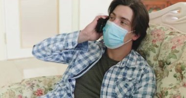 Maskeli bir adam kanepede dinlenirken telefonla konuşuyor.