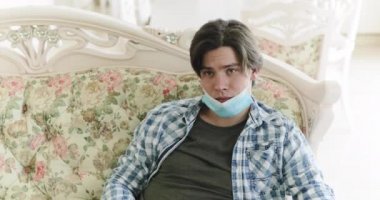 Maskeli çocuk evde kanepede oturuyor, sıkılmış TV seyrediyor ve uzaktan kumanda kullanıyor.