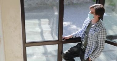 Evdeki güneşli havada sağlık maskeli genç adam pencereden dışarı bakıyor.