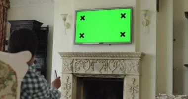 Adam kanepede dinleniyor, yeşil ekranlı TV seyrediyor ve uzaktan kumandayla oynuyor.