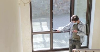 Karantina sırasında pencerenin önünde duran tıbbi maskeli adam akıllı telefon kullanıyor.