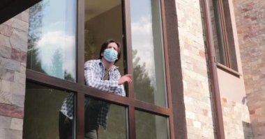 Tıbbi maskeli genç adam açık pencereden bakar ve tecrit hakkında konuşur.