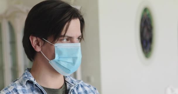 Красивый мужчина в медицинской маске поворачивается и смотрит в камеру на изоляции дома — стоковое видео