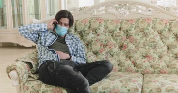 Man med medicinsk mask i ansiktet samtal talar på smartphone när du vilar på soffan — Stockvideo