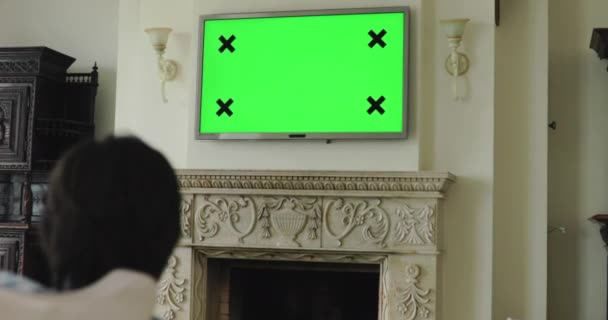 Человек отдыхает на диване, переключает каналы с пульта плазменного телевизора с зеленым экраном — стоковое видео