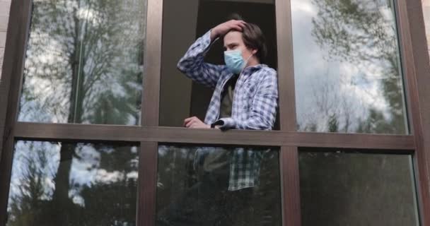 Sad man in medical mask looks through open window and crazes during isolation — Stock Video