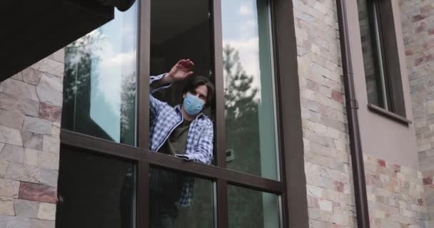 Улыбающийся человек в маске смотрит в открытое окно, машет рукой и показывает палец кому-то — стоковое видео