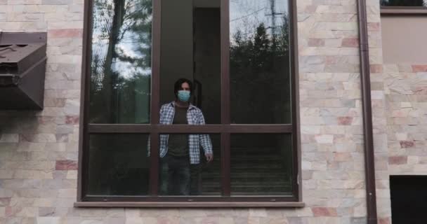 Чоловік у масці приходить до відкритого вікна, спирається на нього і дивиться навколо на відкритому повітрі — стокове відео