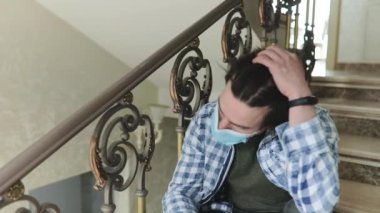 Maskeli adam merdivenlerde tek başına oturuyor, saç stilini düzeltiyor ve düşünüyor.