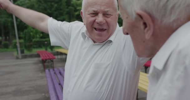 Dva senioři se radují během setkání na lavičce v parku, objímají se a vítají — Stock video