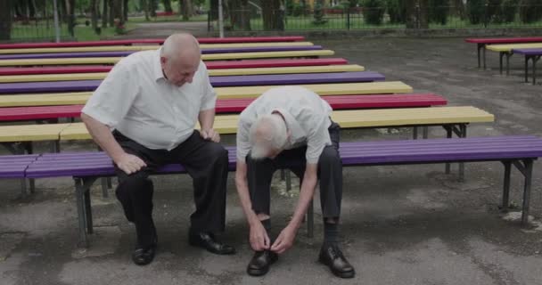 View of two seniors sitting on a park bench and one tying his shoes — 图库视频影像