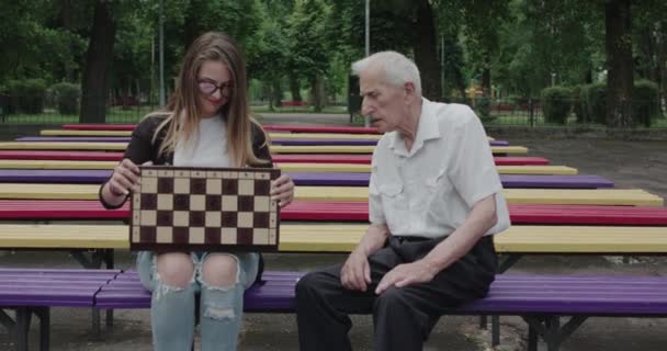 Młoda dziewczyna spędza czas ze staruszkiem w parku pokazuje szachy i śmieje się — Wideo stockowe