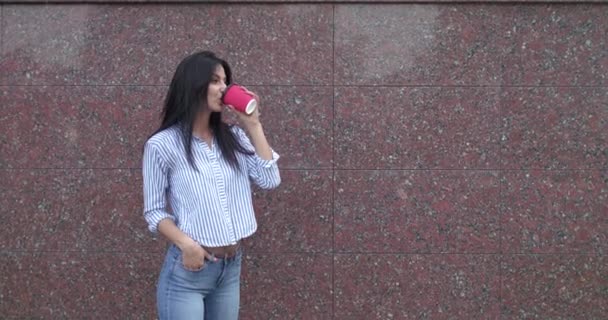Счастливая брюнетка в повседневной одежде пьет чашку напитка и улыбается в камеру — стоковое видео