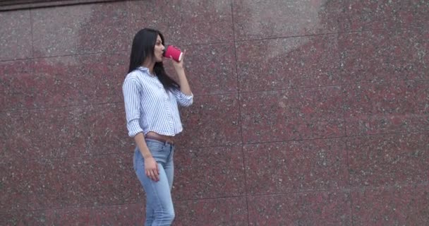 Девушка пьет напиток, кладет руку в карман и смотрит в сторону — стоковое видео
