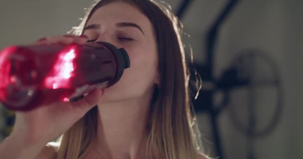 Дівчина п'є зі спортивної пляшки, дивиться на профіль і посміхається на камеру в спортзалі — стокове відео