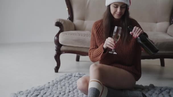 Ładna brunetka w czerwonym kapeluszu relaksuje się na dywanie i nalewa szampana — Wideo stockowe