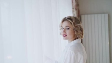 Beyaz gömlekli mutlu kadın pencereye gelir ve kameraya perde atar.