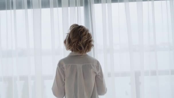 Back look of woman in white shirt comes to light window and scatters curtains — Wideo stockowe