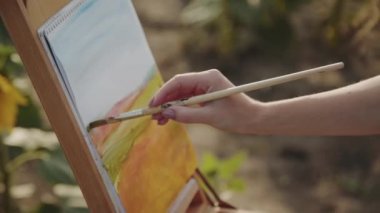 Kadın eli, güneşli doğada suluboya bir manzara çiziyor.