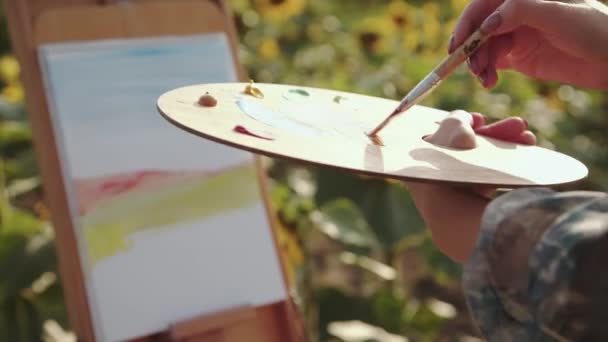 Close view of female hands mix paints with brush on a palette in sunflower field — Videoclip de stoc