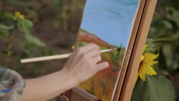 Γυναικείο χέρι ζωγραφίζει ένα τοπίο με πινέλο και ακουαρέλες ανάμεσα σε ηλιοτρόπια — Αρχείο Βίντεο