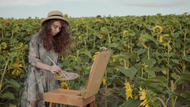 Pretty red-haired lady painting with watercolors among sunflowers — Vídeos de Stock
