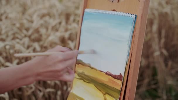 Female hand painting a landscape with brush and watercolors among wheat field — Vídeos de Stock