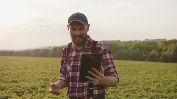 Smiled bearded man talks on tablet on green field — стоковое видео