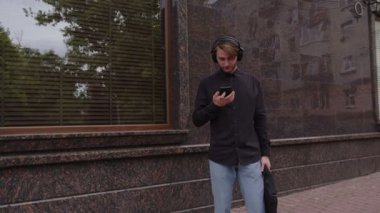 Genç adam kulaklıkla müzik dinliyor, kuryeyle yazıyor ve arkadaşlarını bekliyor.