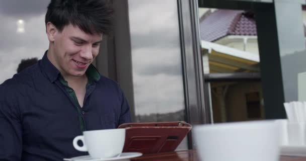 Hombre guapo agita la mano y habla en el chat de vídeo en la tableta en la cafetería al aire libre — Vídeos de Stock