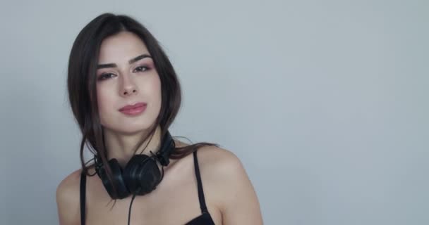 Cute girl with headphones on neck smiles at camera, sways and is ashamed — Stock Video