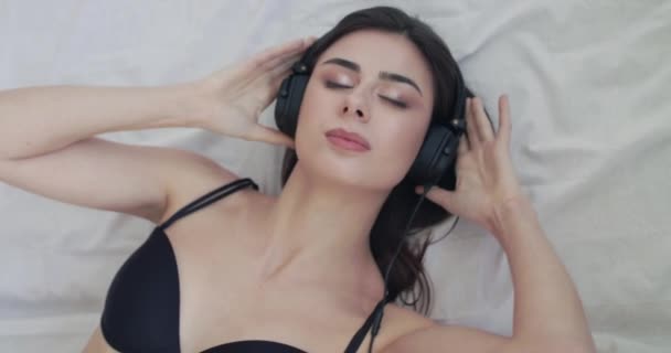 Happy girl berbaring di tempat tidur linen, memakai headphone, bergoyang, melihat ke kamera — Stok Video