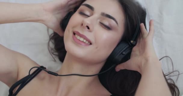 Retrato superior de la mujer acostada en la cama, sostiene los auriculares, balancea y muerde el labio en la cámara — Vídeo de stock