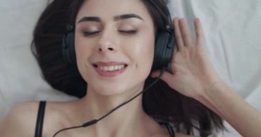 Kadın portresi yatakta uzanıyor, kulaklığı tutuyor, sallanıyor ve kameraya gülümsüyor.