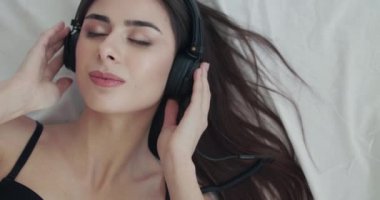 Yatakta müzik dinleyen, kameraya asılan ve kulaklıkları kıvıran bir kız.
